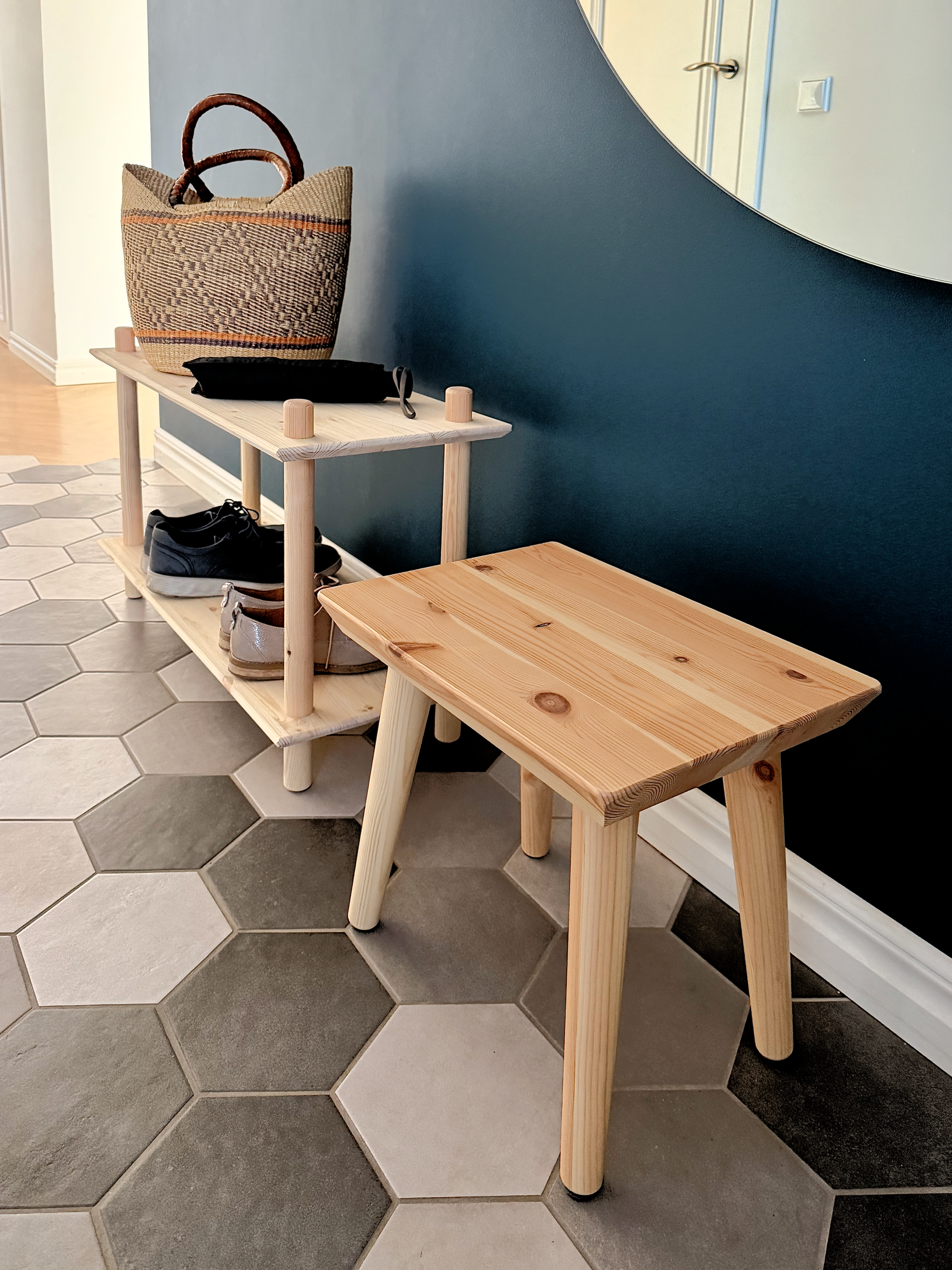 Lifestyle shot featuring the raw pine Air Shelf two-tiered unit holding a woven basket and shoes, placed next to the Air Stool, against a dark blue wall with a round mirror.