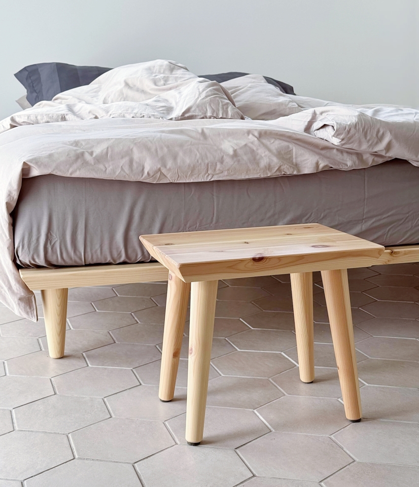 An untreated light pine Air stool positioned next to an Air bed frame 160x200 cm with grey and light bedding, on a hexagonal tiled floor.