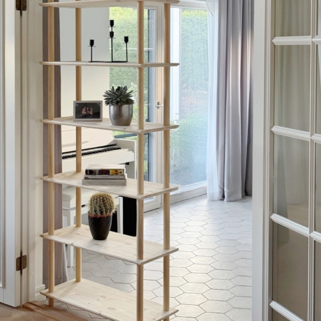 Vue lifestyle de l’étagère Air à six niveaux en pin brut, accueillant une succulente, un cactus, des livres et un cadre photo, placée stratégiquement pour servir de séparateur de pièce dans un espace lumineux et ouvert avec un piano en arrière-plan.