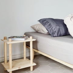 Vue lifestyle montrant l’étagère Rohe à deux niveaux en pin brut utilisée comme table de chevet, placée à côté d’un lit avec des coussins et accueillant une petite pile de livres.