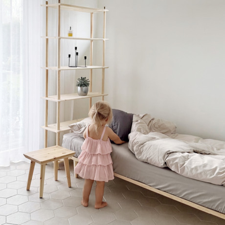 Un petit enfant en robe rose se tient à côté d’un cadre de lit Air 90 × 200 en pin clair non traité, avec un tabouret Air et une grande étagère Air dans une pièce minimaliste.