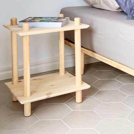 Photo lifestyle montrant l’étagère Rohe à deux niveaux en pin brut utilisée comme table de chevet à côté d’un lit avec des coussins, accueillant une petite pile de livres.