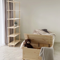 Un coffre Air ouvert en pin non traité, contenant un ours en peluche et une couverture, est placé au pied d’un cadre de lit Air 90 × 200, avec une grande étagère modulaire Air à proximité dans une chambre d’enfant.