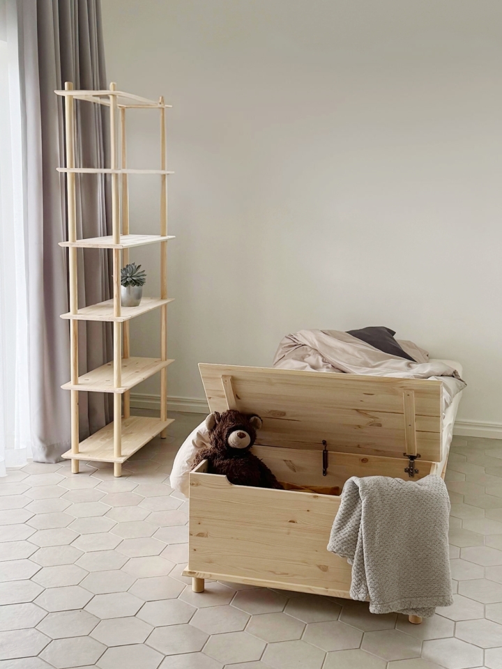 Eine offene Air Truhe aus unbehandeltem Kiefernholz mit einem Teddybär und einer Decke steht am Fußende eines Air Bettgestells 90x200, daneben eine hohe modulare Air Regal-Einheit in einem Kinderzimmer.