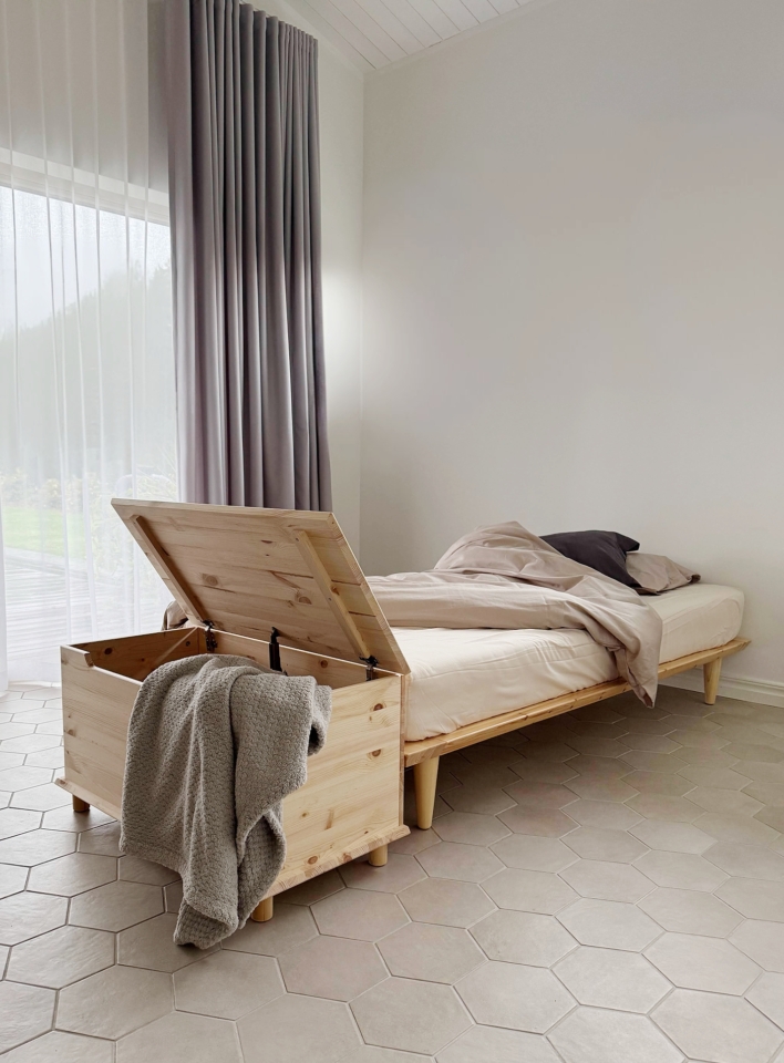 Coffre Air ouvert en pin clair non traité avec une couverture grise, placé à côté d’un cadre de lit Air avec une literie neutre, dans une chambre lumineuse.