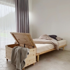 Eine offene Air Truhe aus unbehandeltem hellem Kiefernholz mit einer grauen Decke, neben einem Air Bettgestell mit neutraler Bettwäsche, in einem hellen Schlafzimmer positioniert.