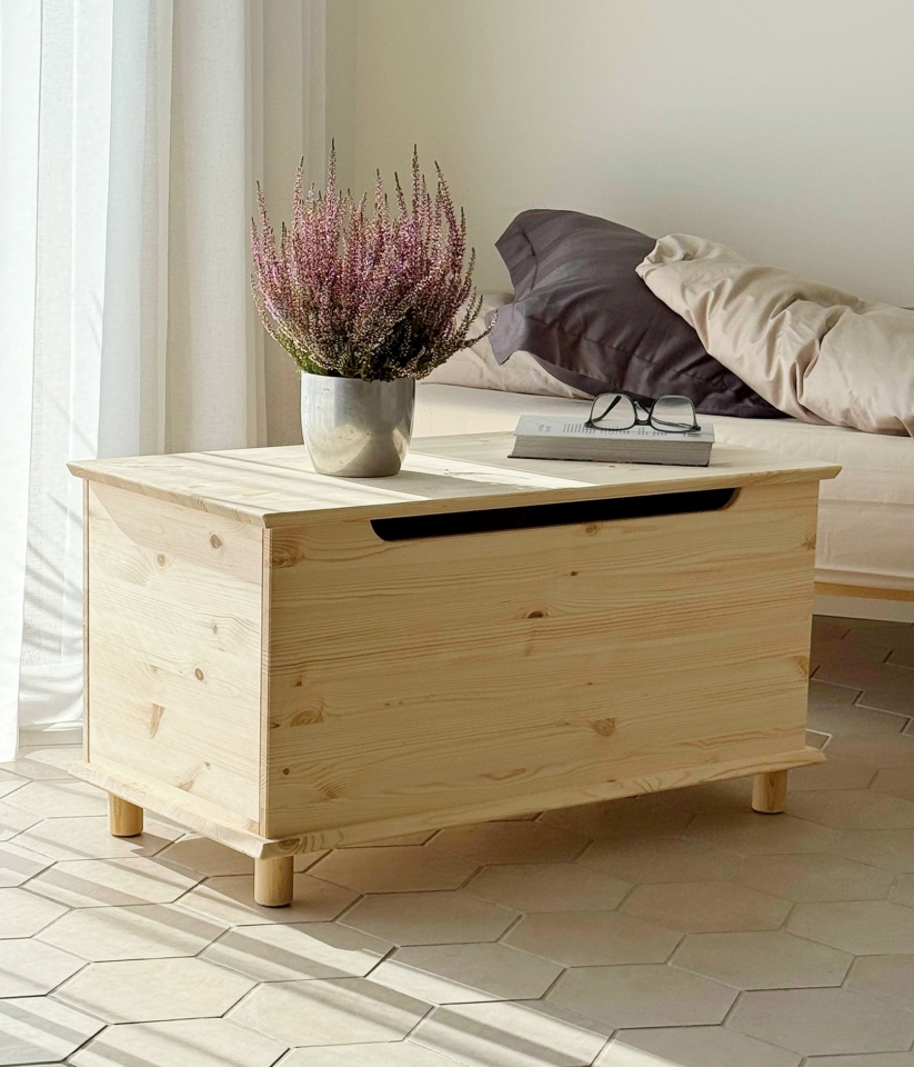 Coffre Air en pin clair non traité, avec une plante en pot et des lunettes posées dessus, placé à côté d’un cadre de lit Air avec une literie neutre, sur un sol carrelé.