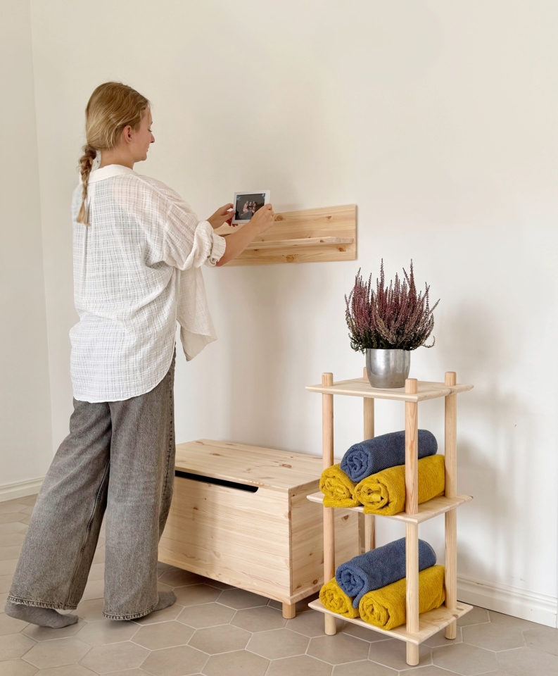 Une femme place une tablette numérique sur une étagère murale Air en pin non traité. En dessous se trouvent un coffre Air et une étagère Air étroite avec des serviettes roulées et une plante, dans une pièce lumineuse.
