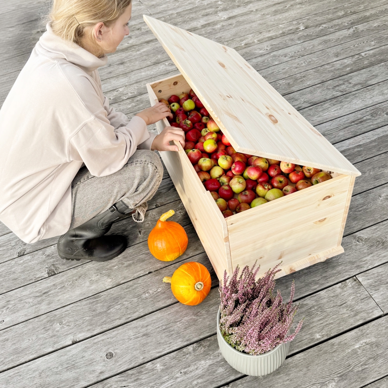 Une jeune femme plonge la main dans un coffre Air ouvert en pin clair non traité, rempli de pommes rouges et vertes, posé sur une terrasse en bois avec des citrouilles et une plante à proximité.