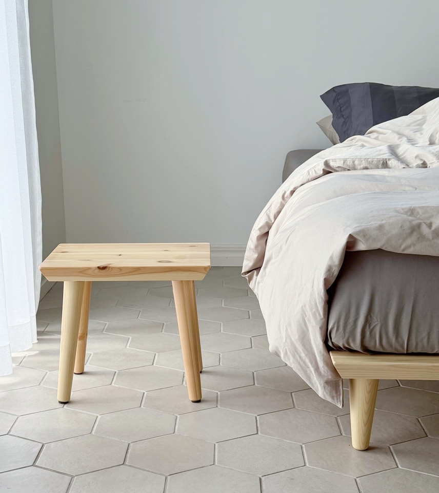 Tabouret Air en pin clair non traité posé sur un sol carrelé hexagonal clair, placé à côté d’un lit Air avec une literie neutre, vu depuis un angle bas.