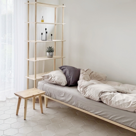Ein lichtdurchflutetes Schlafzimmer mit einem schlichten Holzbett, einem Air Hocker aus unbehandeltem Kiefernholz und einer hohen Air Regal-Einheit aus Holz, alles Teil einer minimalistischen Ästhetik auf einem gefliesten Boden.