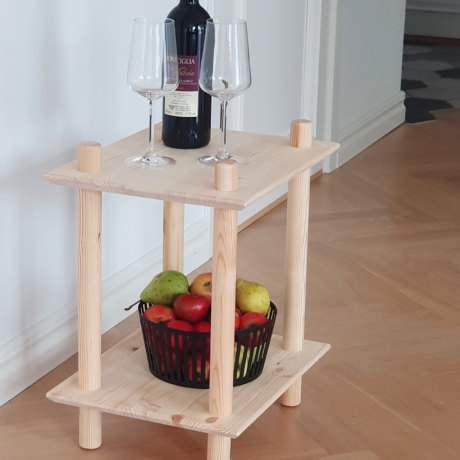 Vue lifestyle en gros plan de l’étagère modulaire à deux niveaux en pin brut, présentant une bouteille de vin rouge et deux verres sur l’étagère supérieure, ainsi qu’un panier noir rempli de pommes et de poires sur l’étagère inférieure.