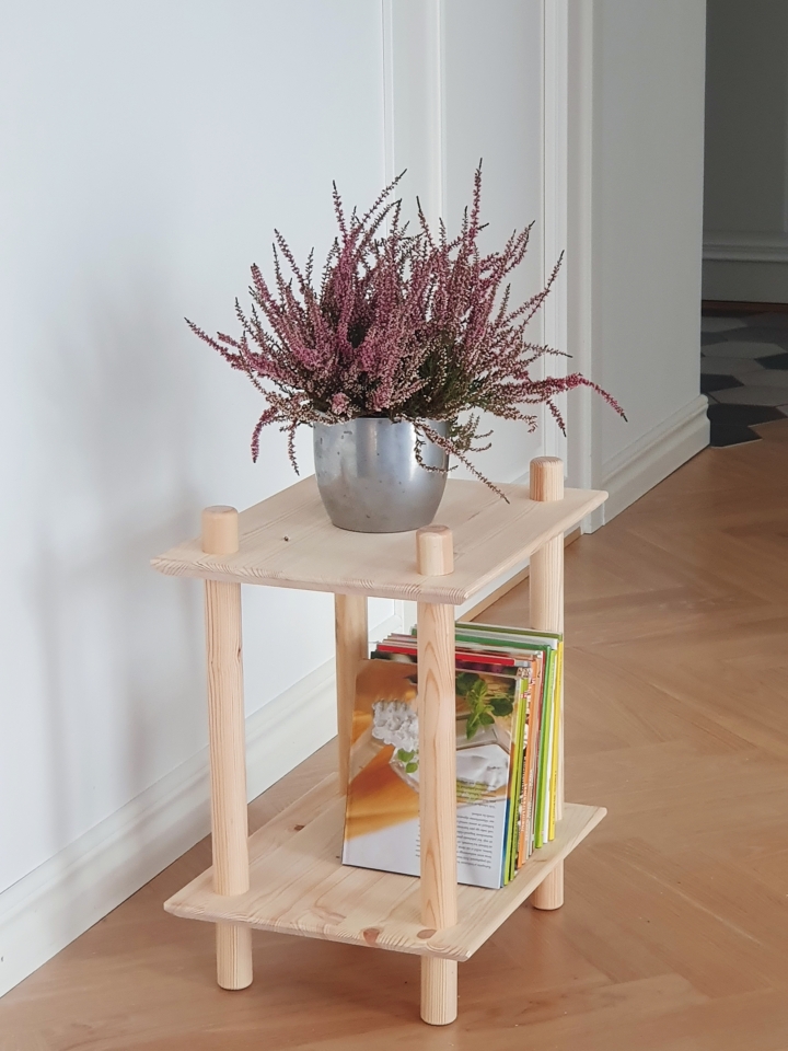 Vue lifestyle de l’étagère modulaire à deux niveaux en pin brut, placée dans un coin de la pièce, mise en scène avec un pot argenté de bruyère violette et une pile de livres colorés sur l’étagère inférieure.