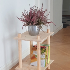 Lifestyleaufnahme des zweistufigen modularen Regals aus rohem Kiefernholz in einer Raumecke, gestaltet mit einem silbernen Topf mit violetter Heide und einem Stapel farbenfroher Bücher auf der unteren Ablage.