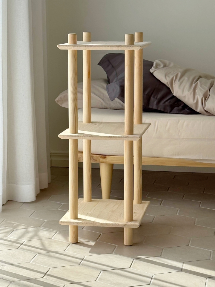 Vue lifestyle de l’étagère Air étroite modulaire à trois niveaux en pin brut, placée dans un coin de chambre, à côté d’un lit avec des coussins, éclairée par la lumière naturelle.
