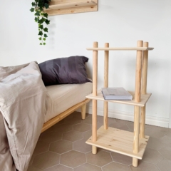 Vue lifestyle de l’étagère Air étroite modulaire à trois niveaux en pin brut, placée à côté d’un lit, accueillant un livre, avec une étagère murale assortie au-dessus, mise en scène avec une plante retombante de lierre.