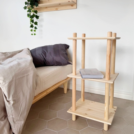 Lifestyleaufnahme der schmalen dreistufigen modularen Air Shelf Einheit aus rohem Kiefernholz neben einem Bett, die ein Buch hält, mit einem passenden Wandregal darüber, dekoriert mit einer herabhängenden Efeupflanze.