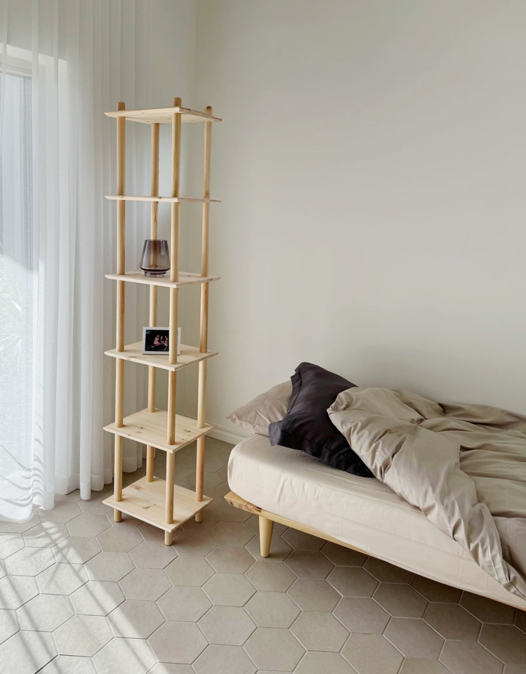 Étagère modulaire Air haute en pin clair non traité avec un vase et un cadre photo, placée à côté d’un cadre de lit Air avec une literie neutre, dans une pièce minimaliste baignée de lumière naturelle.