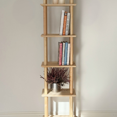 Vue lifestyle en pleine hauteur de l’étagère Air étroite à six niveaux en pin brut, accueillant des piles de livres colorés et un pot argenté avec de la bruyère violette, placée contre un mur blanc et un sol en bois clair.