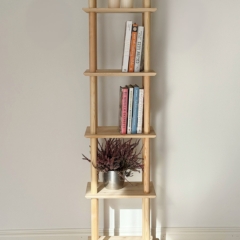 Ganzkörper-Lifestyleaufnahme der schmalen sechsstufigen Air Shelf Einheit aus rohem Kiefernholz, die Stapel farbenfroher Bücher und einen silbernen Topf mit violetter Heide enthält, platziert vor einer weißen Wand und einem hellen Holzboden.