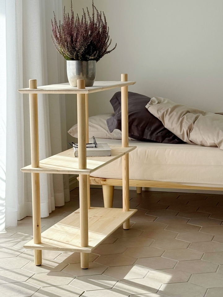 Étagère Air étroite en pin clair non traité, avec une plante en pot et des livres, placée à côté d’un cadre de lit Air avec literie, sur un sol carrelé baigné de lumière naturelle.