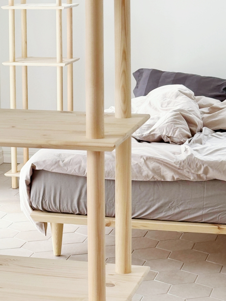 Detail-Nahaufnahme des sechsstöckigen modularen Air Regals aus unbehandeltem Kiefernholz, die die Holzpfosten und Regalböden im Vordergrund zeigt, mit einem minimalistischen Bett und grauer Bettwäsche im Hintergrund.