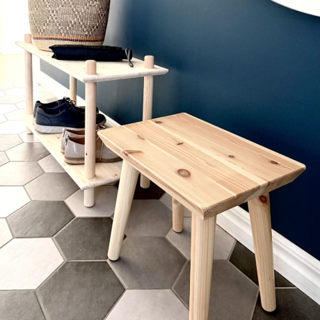 Un tabouret Air en pin clair non traité est posé sur un sol carrelé à motifs hexagonaux, placé devant une étagère Air contre un mur bleu foncé.