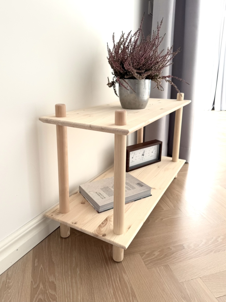 Photo lifestyle de l’étagère modulaire Air large à deux niveaux en pin brut, mise en scène avec une plante de bruyère violette en pot, un livre et une horloge de style vintage, placée contre un mur blanc et sur un sol en bois.