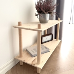 Photo lifestyle de l’étagère modulaire Air large à deux niveaux en pin brut, mise en scène avec une plante de bruyère violette en pot, un livre et une horloge de style vintage, placée contre un mur blanc et sur un sol en bois.