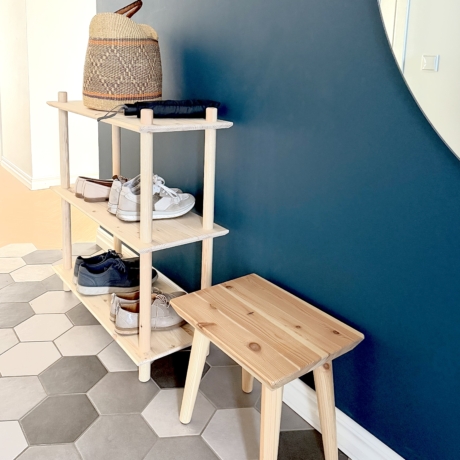 Lifestyleaufnahme mit dem dreistöckigen Air Regal aus unbehandeltem Kiefernholz, das mehrere Paar Schuhe trägt, platziert neben dem Air Hocker, vor einer dunkelblauen Wand und auf einem gemusterten Boden im Eingangsbereich.