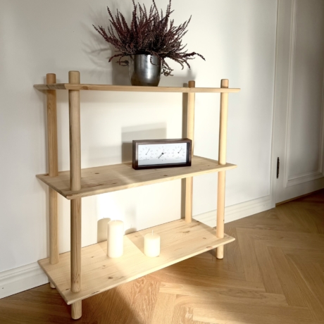 Photo lifestyle de l’étagère Air large à trois niveaux en pin brut, mise en scène avec une plante de bruyère en pot sur l’étagère supérieure, une horloge en bois sur l’étagère centrale et deux bougies cylindriques blanches sur l’étagère inférieure.