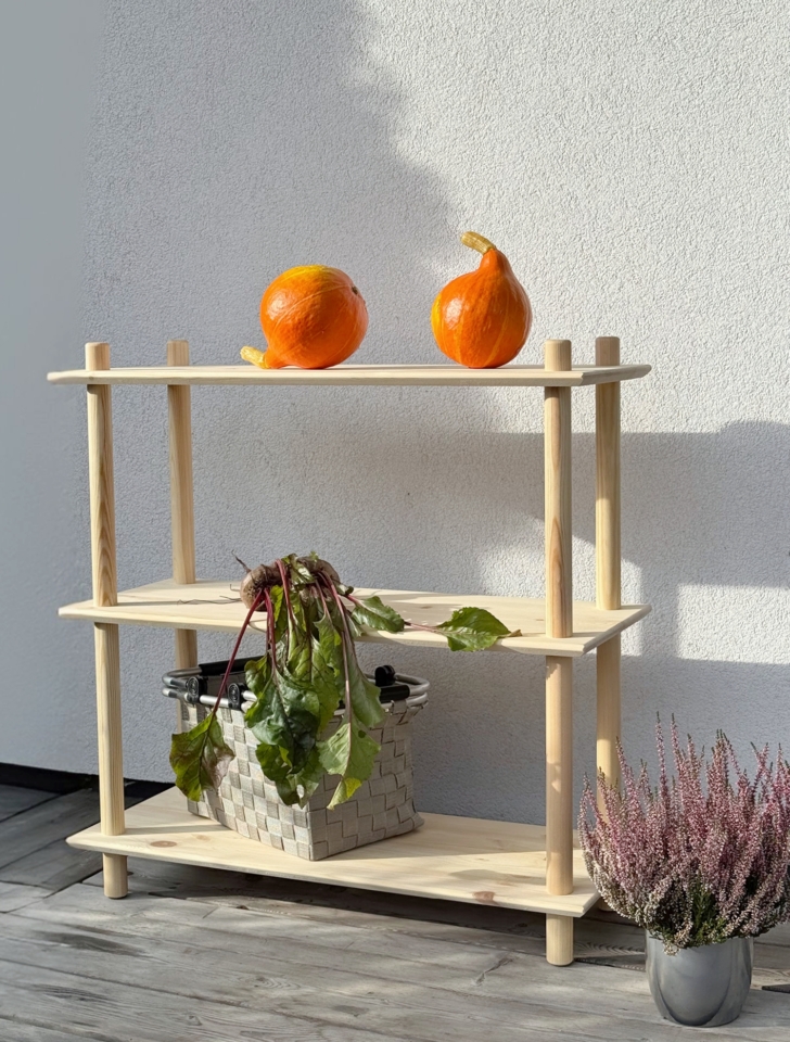 Photo lifestyle en extérieur de l’étagère Air large à trois niveaux en pin brut, mettant en scène deux citrouilles orange sur l’étagère supérieure et un panier de betteraves fraîches sur l’étagère intermédiaire, à côté d’une plante de bruyère en pot.