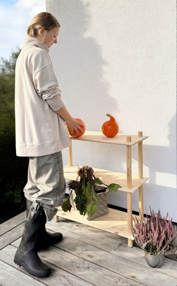 Une femme portant un sweat clair et des bottes dépose une citrouille orange sur l’étagère supérieure d’une étagère Air large à trois niveaux en pin brut, qui accueille également un panier de betteraves, sur une terrasse en bois.