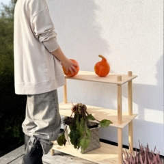 Une femme portant un sweat clair et des bottes dépose une citrouille orange sur l’étagère supérieure d’une étagère Air large à trois niveaux en pin brut, qui accueille également un panier de betteraves, sur une terrasse en bois.