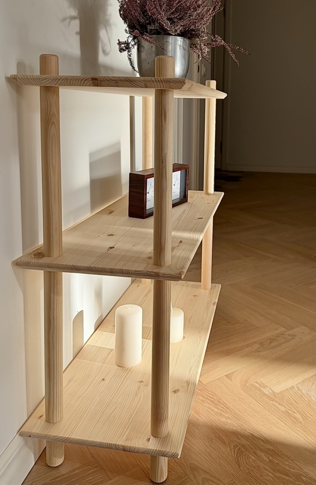 Lifestyleaufnahme des breiten, zweistöckigen modularen Air Regals aus unbehandeltem Kiefernholz, gestaltet mit einer violetten Heidepflanze im Topf auf der oberen Ablage, einer dekorativen Holzuhr und einem Buch auf dem unteren Regal, vor einer weißen Wand.