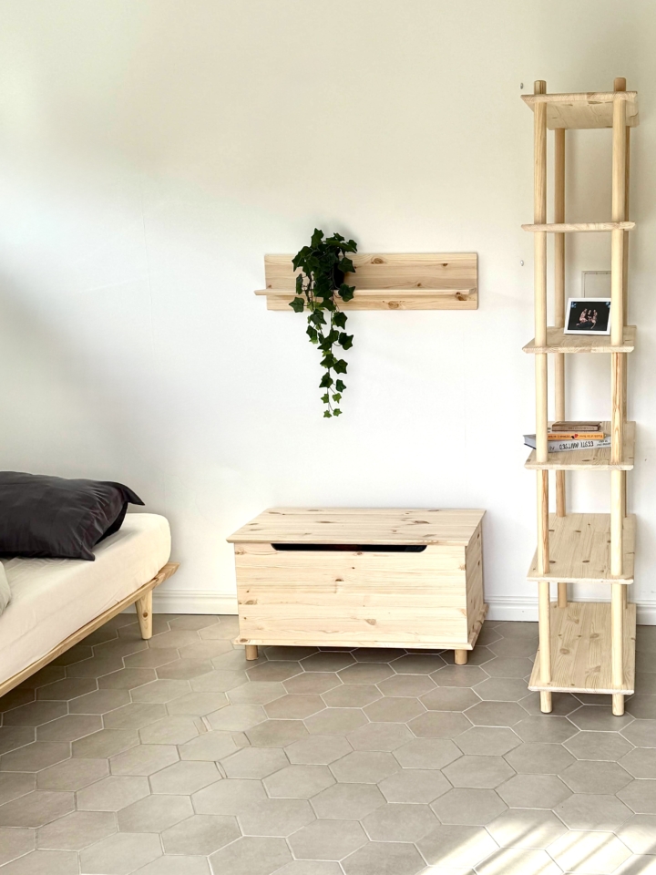 Ganzaufnahme des Raumes mit dem schmalen, sechsstöckigen Air Regal aus unbehandeltem Kiefernholz, einem passenden Wandregal mit Efeu, einer Holz-Aufbewahrungsbank und einem Bett mit grauer Bettwäsche – alles aus derselben Kollektion.