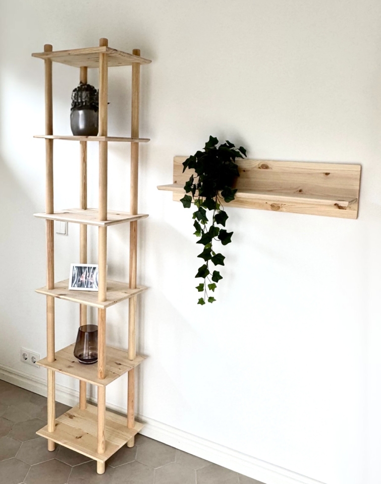 Photo lifestyle mettant en scène la grande étagère Air à six niveaux en pin brut, accueillant une plante, une photo encadrée et un vase, aux côtés d’une étagère murale Air assortie avec une plante retombante de lierre, le tout contre un mur blanc.