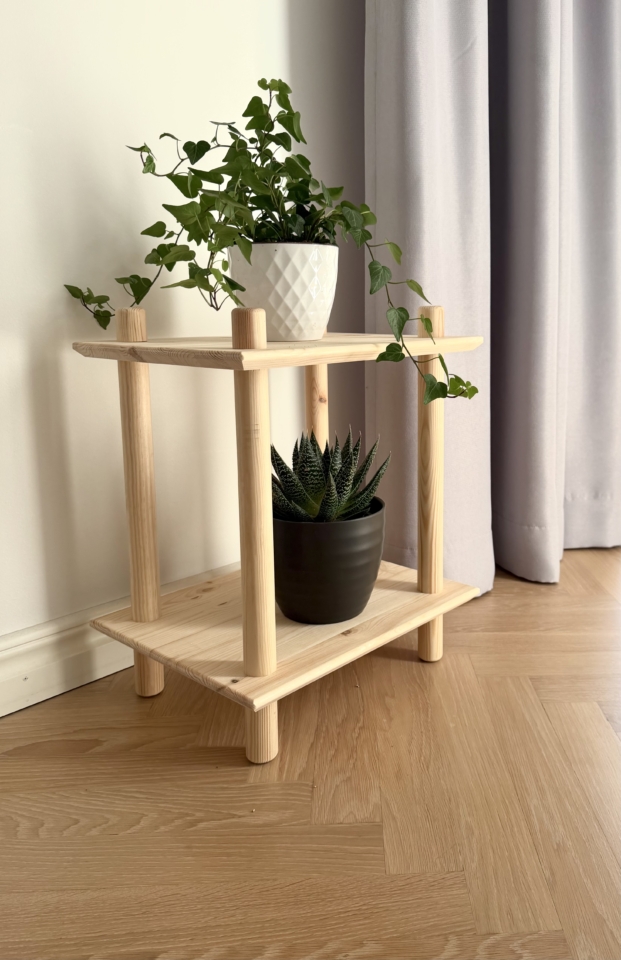 Lifestyleaufnahme des zweistöckigen modularen Regals aus unbehandeltem Kiefernholz vor einer weißen Wand und einem grauen Vorhang, gestaltet mit zwei Topf-Zimmerpflanzen (Sukkulente und Efeu).