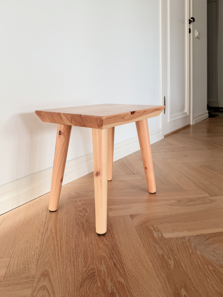 Un tabouret Air en pin clair non traité est posé sur un parquet à chevrons dans une entrée lumineuse, avec un mur blanc et une porte fermée en arrière-plan.
