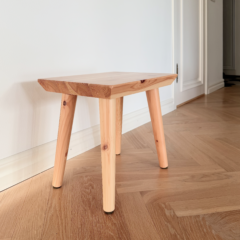 Un tabouret Air en pin clair non traité est posé sur un parquet à chevrons dans une entrée lumineuse, avec un mur blanc et une porte fermée en arrière-plan.
