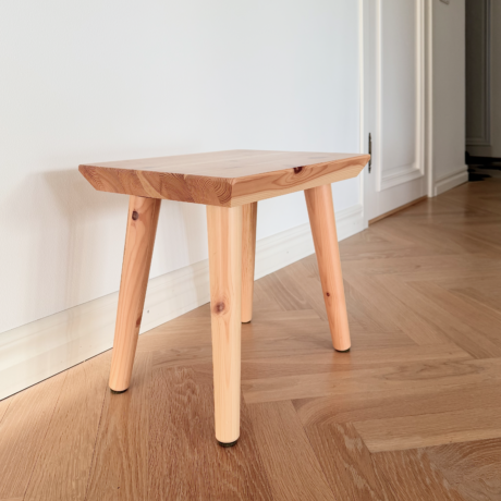 Un tabouret Air en pin clair non traité est posé sur un parquet à chevrons dans une entrée lumineuse, avec un mur blanc et une porte fermée en arrière-plan.