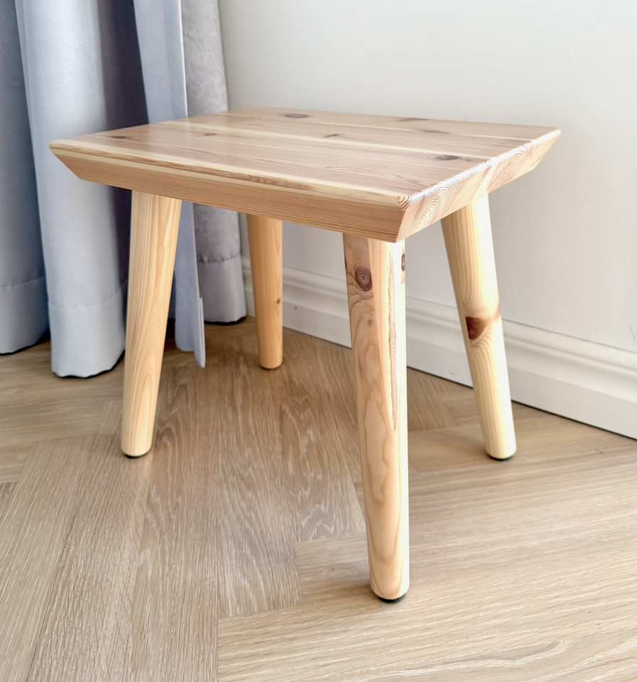 Gros plan du tabouret Air en pin clair non traité sur un sol en bois clair, mettant en valeur le veinage naturel et les nœuds subtils, avec un rideau en arrière-plan.