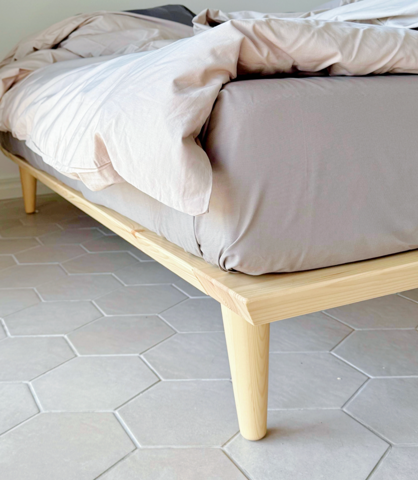 Nahaufnahme des konisch zulaufenden Holzbeins und der Rahmenecke eines Air Bettgestells 180x200 cm aus unbehandeltem, hellem Kiefernholz mit Bettwäsche auf einem sechseckigen Fliesenboden.