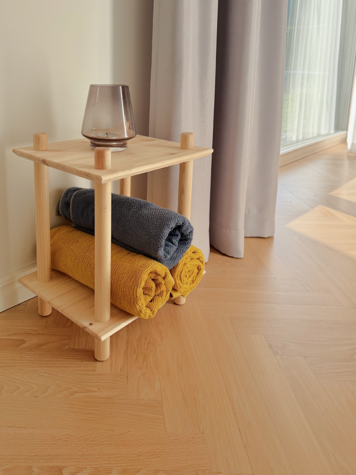 Photo lifestyle de l’étagère modulaire Air à deux niveaux en pin brut, placée contre un mur clair et un rideau léger, mise en scène avec deux serviettes roulées (bleue et jaune) et un petit vase en verre sur le dessus.