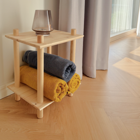 Photo lifestyle de l’étagère modulaire Air à deux niveaux en pin brut, placée contre un mur clair et un rideau léger, mise en scène avec deux serviettes roulées (bleue et jaune) et un petit vase en verre sur le dessus.