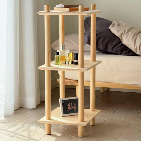 Lifestyle-Nahaufnahme des schmalen, dreistöckigen Air Regals aus unbehandeltem Kiefernholz mit verschiedenen Parfümflakons und Lippenstiften auf der oberen Ablage sowie einem gerahmten Bild auf der unteren Ablage neben einem Bett.