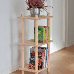 Lifestyleaufnahme des schmalen, dreistöckigen Air Regals aus unbehandeltem Kiefernholz, gestaltet mit einem silbernen Topf mit violetter Heide auf der oberen Ablage sowie mehreren farbenfrohen Büchern und Kochbüchern auf den beiden unteren Regalböden.