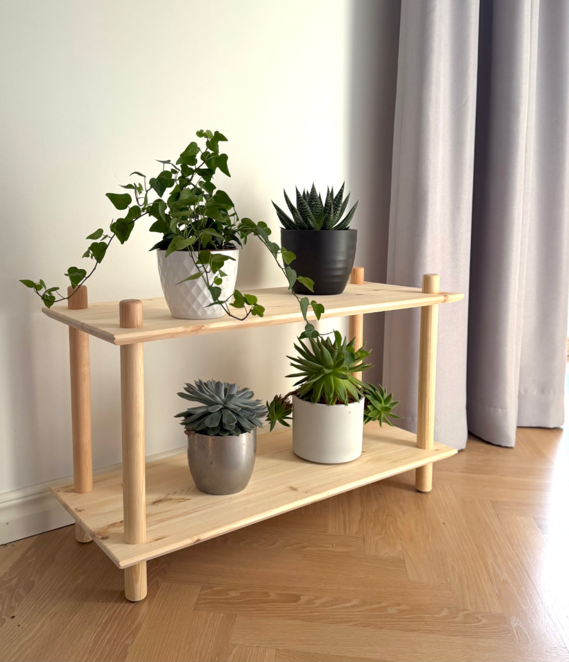 Photo lifestyle de l’étagère modulaire Air large à deux niveaux en pin brut, garnie de quatre plantes d’intérieur en pot (lierre, succulentes), contre un mur blanc et un rideau gris, sur un sol en bois clair.