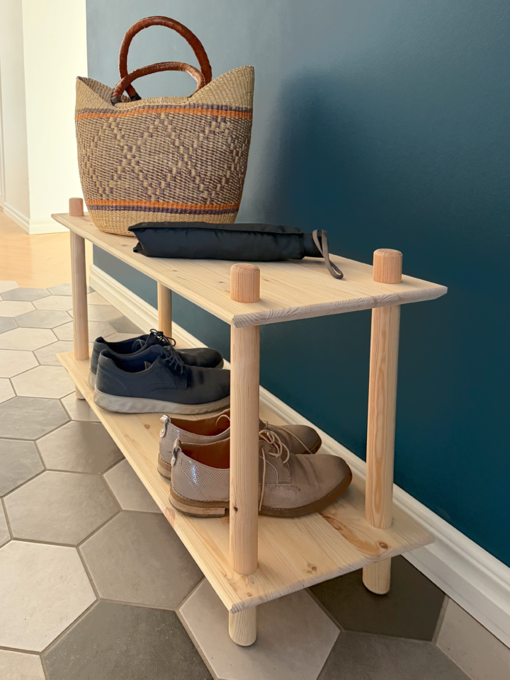 Photo lifestyle de l’étagère Air large à deux niveaux en pin brut dans une entrée, accueillant un panier tressé et un parapluie sur le niveau supérieur, avec deux paires de chaussures rangées sur l’étagère inférieure.