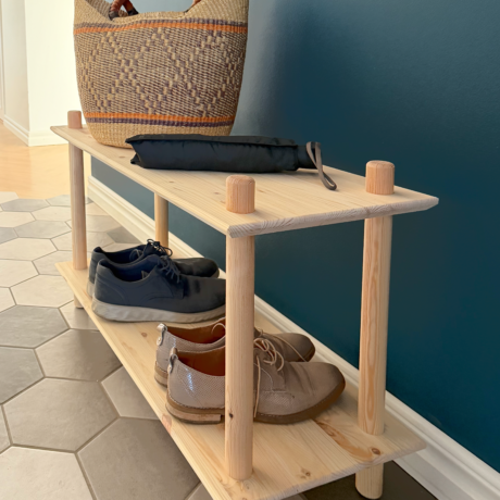Photo lifestyle de l’étagère Air large à deux niveaux en pin brut dans une entrée, accueillant un panier tressé et un parapluie sur le niveau supérieur, avec deux paires de chaussures rangées sur l’étagère inférieure.